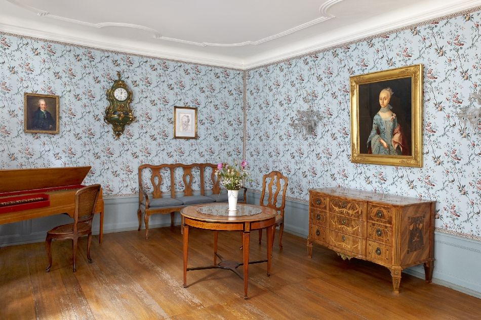 Zimmer mit antiken M&ouml;beln, blau-gebl&uuml;mter Tapete und Gem&auml;lden und Uhr an der Wand