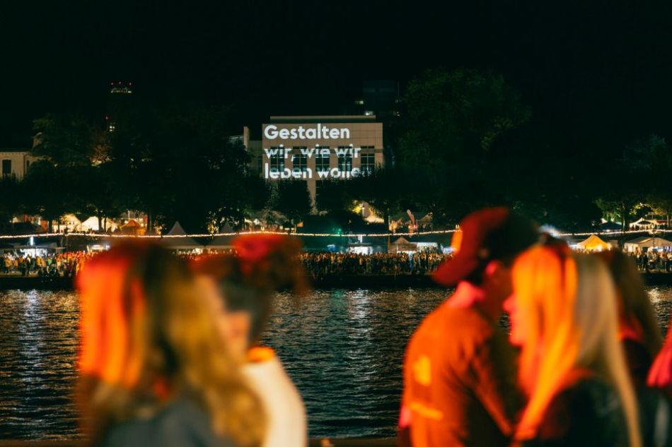 Menschen spazieren bei Nacht am Flussufer, auf ein Haus ist 'Gestalten wir, wie wir leben wollen' projiziert