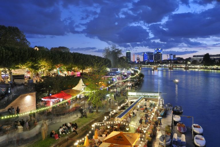 Buden und Bühnen mit erleuchtetem Boot am Main bei Nacht und Besucher*innen des Museumsuferfests